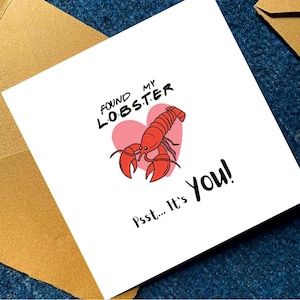 Pu&ograve; includere: Un biglietto d'auguri quadrato bianco con un'illustrazione di un'aragosta rossa centrata sopra un cuore rosa. Il testo recita "FOUND MY LOBSTER" in lettere nere con punti colorati e "Psst... It's YOU!" in corsivo nero. Accompagnato da buste marroni.