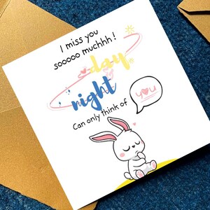 Op de afbeelding: Een vierkante wenskaart met een getekend wit konijn met roze oren en pootjes. De tekst luidt: I miss you sooooo muchhh! day night can only think of you. Het ontwerp bevat een gele zon, een roze orbitale lijn en een tekstballon met het woord you.
