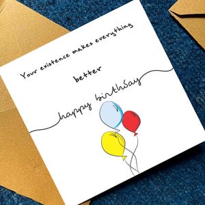 May include: A white birthday card with the message "Your existence makes everything better" and "happy birthday" written in black script. The card features three balloons in blue, red, and yellow. The card is placed on a blue textured surface with a gold envelope.