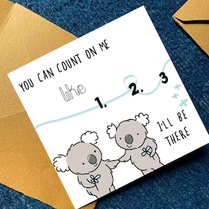 May include: A white greeting card with the text "You can count on me like 1. 2. 3. I'll be there." features two cartoon koalas holding hands. The card is on a blue surface with a brown envelope.