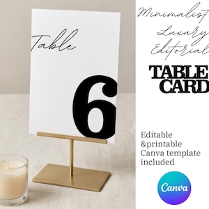 Op de afbeelding: Een witte tafelkaart met het zwarte nummer 6, weergegeven in een gouden metalen standaard. Het woord "Table" is geschreven in een scriptlettertype. De woorden "TABLE CARD" staan vetgedrukt in zwarte letters.