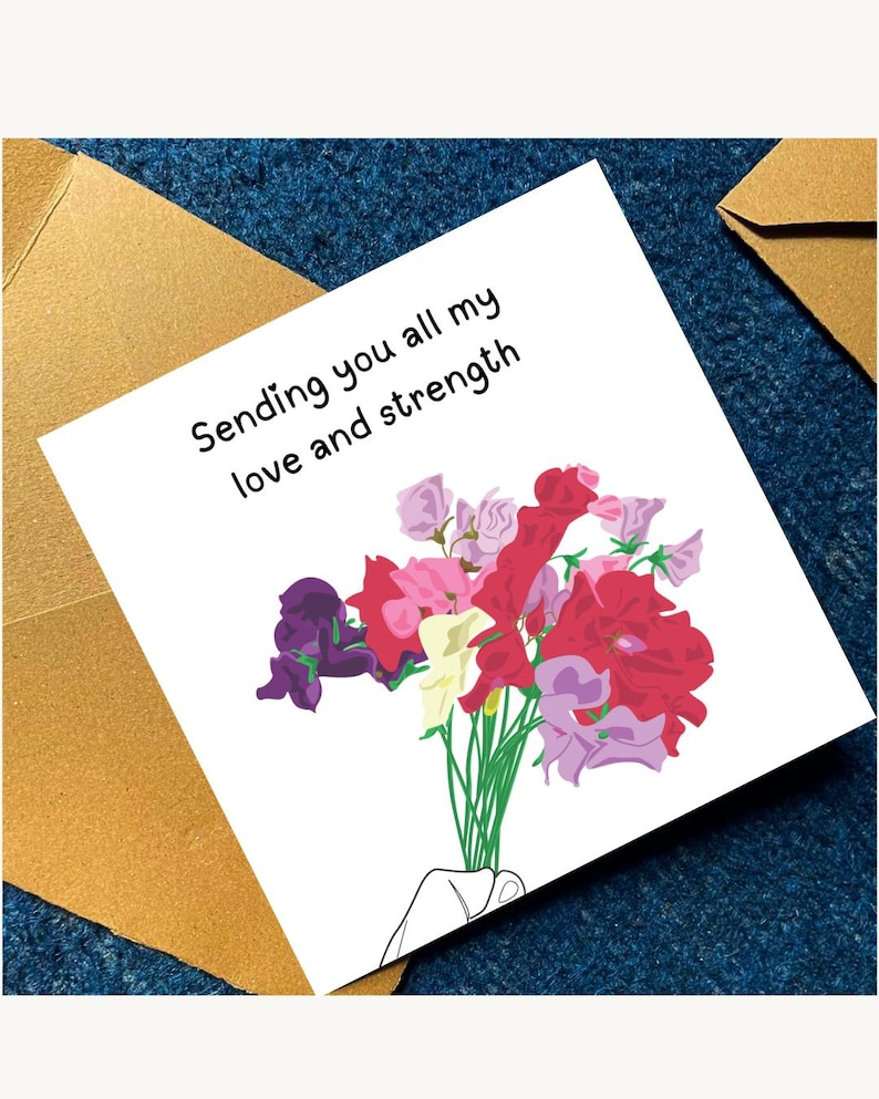 May include: A greeting card with the message "Sending you all my love and strength" and a bouquet of flowers in shades of red, pink, purple, and yellow. The card is on a blue textured surface, with a gold envelope in the background.