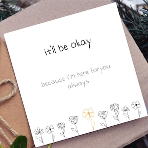 May include: A white card with the text "it'll be okay" and "because I'm here for you always". The card features a line drawing of flowers at the bottom, with one flower in gold. The card is on a brown surface.
