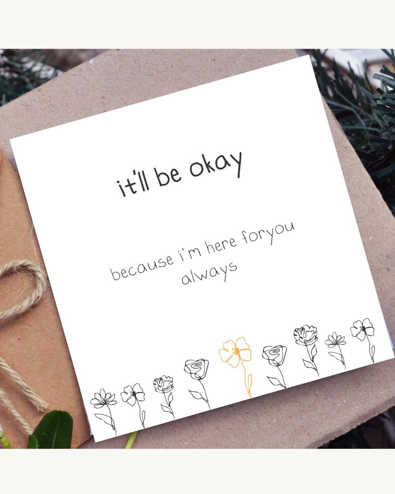 May include: A white card with the text "it'll be okay" and "because I'm here for you always". The card features a line drawing of flowers at the bottom, with one flower in gold. The card is on a brown surface.