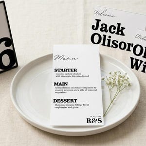 May include: A wedding table setting with a menu, place cards, and table number. The menu lists starter, main, and dessert options. Place cards read "Welcome Jack Olisor" and "Welcome Olivia Wilson." Table number 6 is displayed.