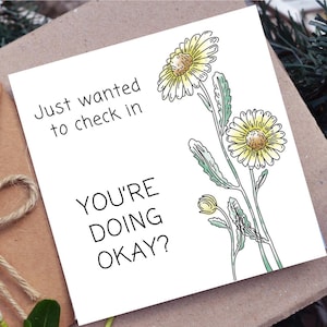 May include: White greeting card with the text "Just wanted to check in, YOU'RE DOING OKAY?" and a watercolor illustration of yellow daisies with green leaves. The card is on a brown surface.