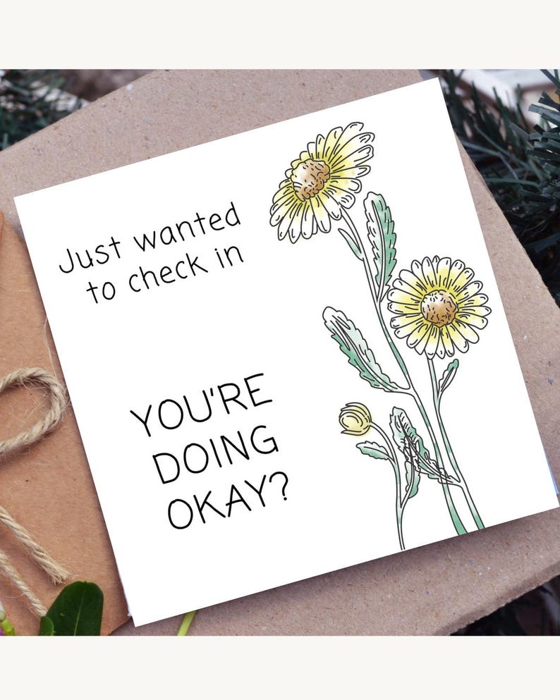 May include: White greeting card with the text "Just wanted to check in, YOU'RE DOING OKAY?" and a watercolor illustration of yellow daisies with green leaves. The card is on a brown surface.