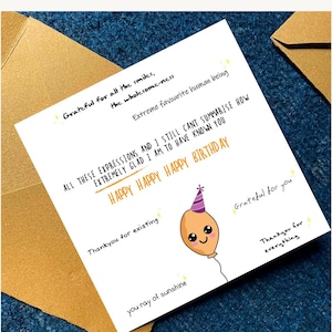 May include: A white birthday card with handwritten text and a cartoon balloon illustration. The card includes phrases like "Happy Birthday," "Grateful for you," and "You ray of sunshine." The card is placed on a blue surface.