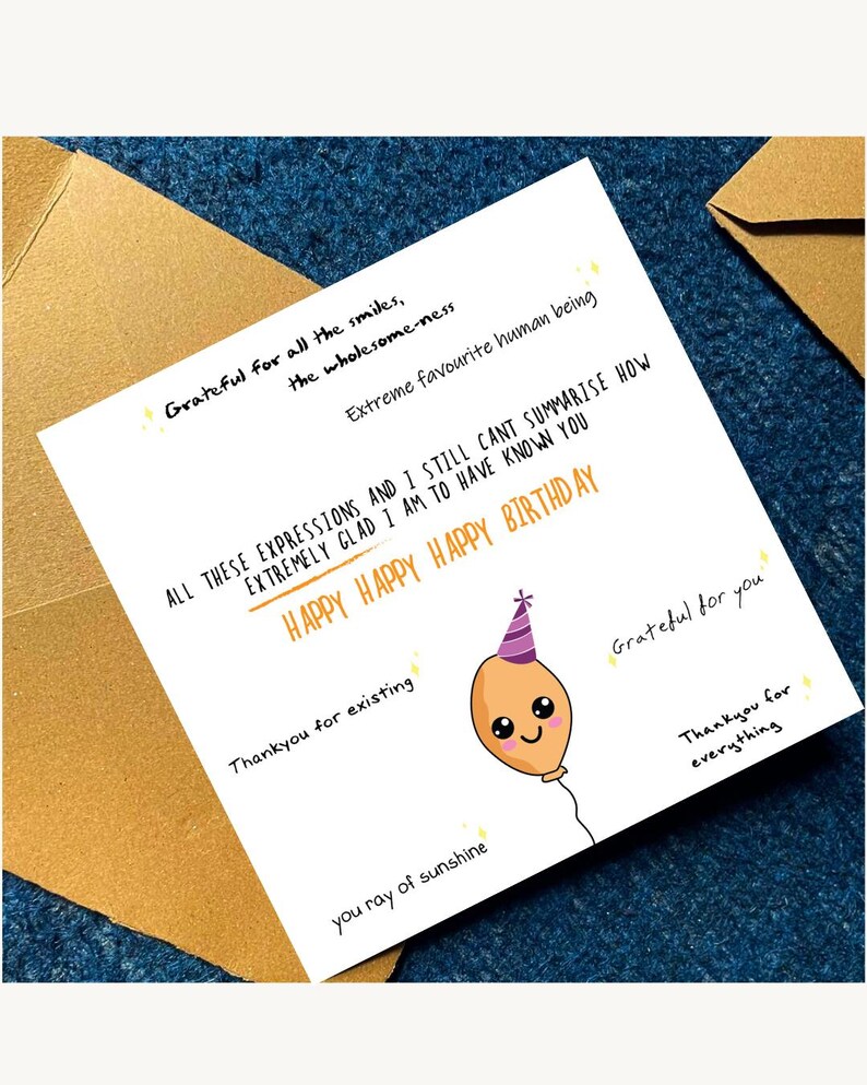May include: A white birthday card with handwritten text and a cartoon balloon illustration. The card includes phrases like "Happy Birthday," "Grateful for you," and "You ray of sunshine." The card is placed on a blue surface.