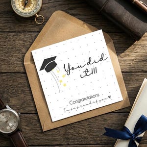 Peut inclure: Une carte de v&oelig;ux carr&eacute;e blanche avec une ic&ocirc;ne de toque de dipl&ocirc;m&eacute; noire, des &eacute;toiles jaunes et le texte "You did it!!!", "Congratulations" et "I'm so proud of you" avec un petit c&oelig;ur, gliss&eacute;e dans une enveloppe brune sur une surface en bois.