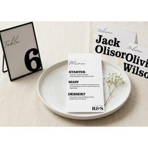 Op de afbeelding: Een bruiloftstafel met een wit bord, menu en tafelkaarten. Het menu bevat een voorgerecht, hoofdgerecht en dessert. Een tafelkaart met nummer 6 staat in een zwart frame. De tafelkaarten zeggen "Welkom".