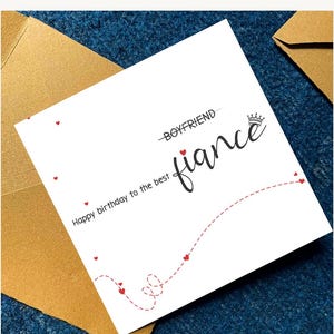 Puede incluir: Una tarjeta de cumplea&ntilde;os blanca con la palabra "fianc&eacute;" en cursiva, una corona y el texto "Happy birthday to the best". Peque&ntilde;os corazones rojos y una l&iacute;nea punteada decoran la tarjeta. La tarjeta est&aacute; colocada sobre una superficie azul con un sobre dorado.