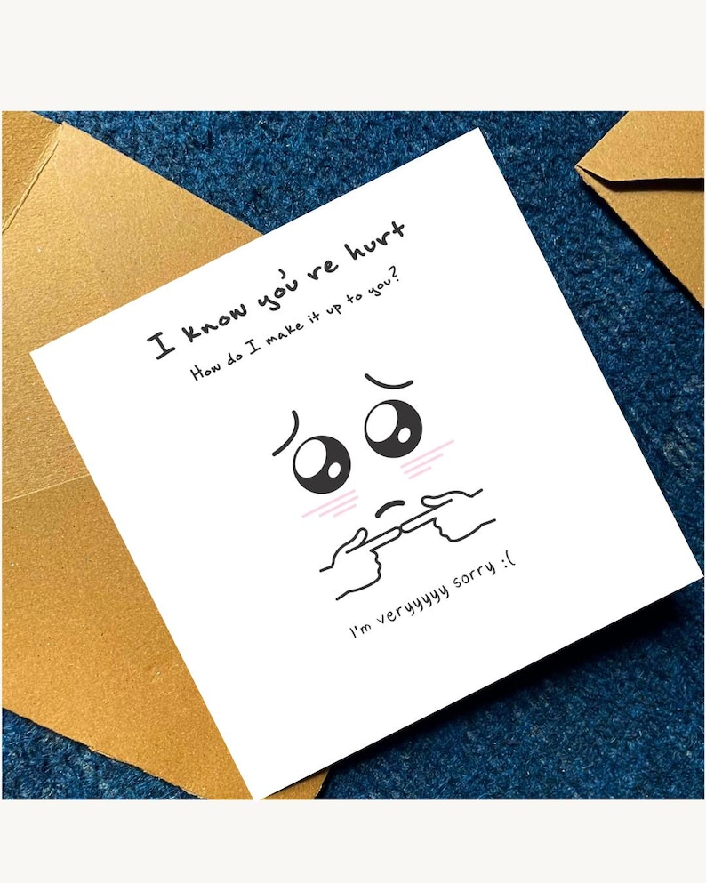 May include: A white greeting card with the text "I know you're hurt. How do I make it up to you?" and "I'm veryyyyy sorry :(". The card features a cartoon face with sad eyes and hands pointing to the mouth. The card is placed on a blue surface with a brown envelope.