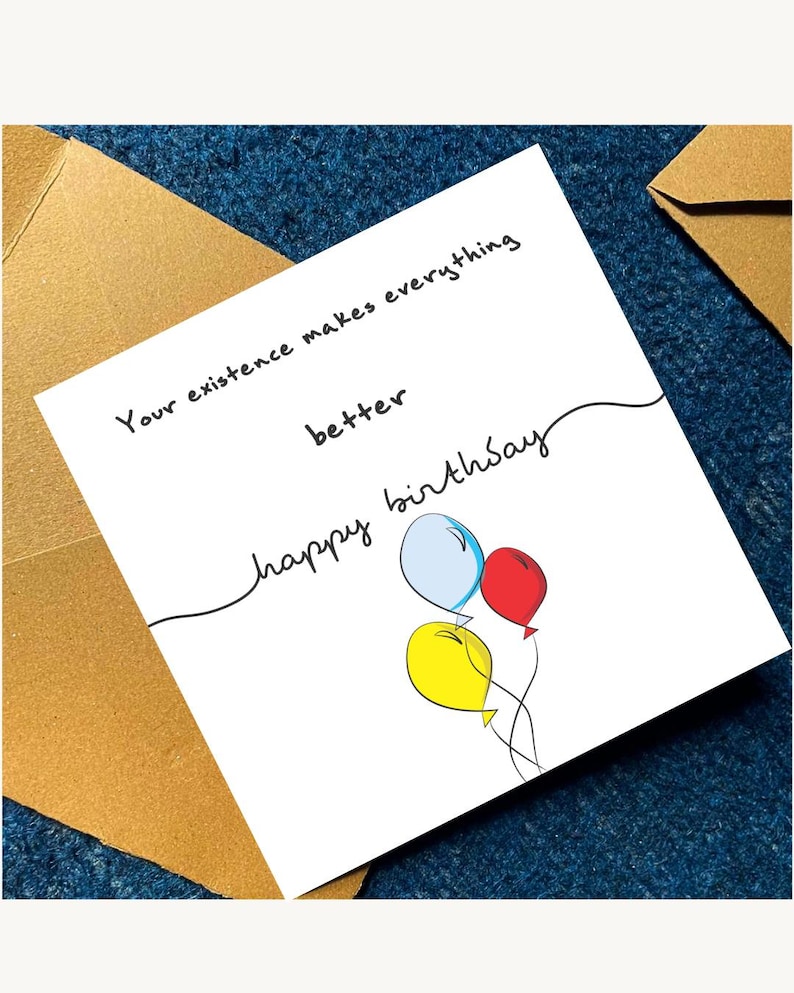 May include: A white birthday card with the message "Your existence makes everything better" and "happy birthday" written in black script. The card features three balloons in blue, red, and yellow. The card is placed on a blue textured surface with a gold envelope.