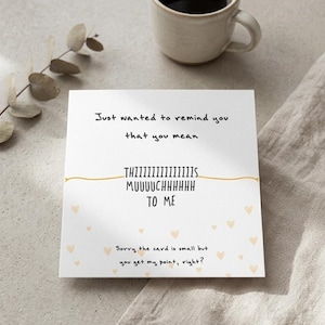 Puede incluir: Una tarjeta blanca con el texto "Just wanted to remind you that you mean THIIIIIIIIIIIIIIS MUUUUCHHHHHH TO ME." La tarjeta tiene peque&ntilde;as ilustraciones de corazones y est&aacute; junto a una taza beige y una ramita.