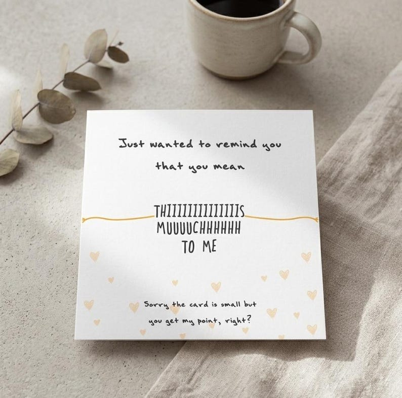 Puede incluir: Una tarjeta blanca con el texto "Just wanted to remind you that you mean THIIIIIIIIIIIIIIS MUUUUCHHHHHH TO ME." La tarjeta tiene peque&ntilde;as ilustraciones de corazones y est&aacute; junto a una taza beige y una ramita.