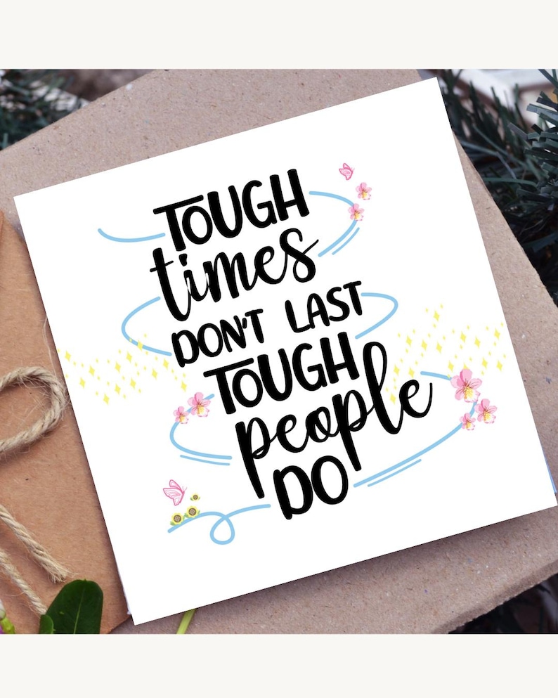 May include: A white greeting card with the text "TOUGH times DON'T LAST TOUGH people DO" in black script. The card is decorated with light blue swirls, pink flowers, and yellow starbursts. The card is on a brown surface.