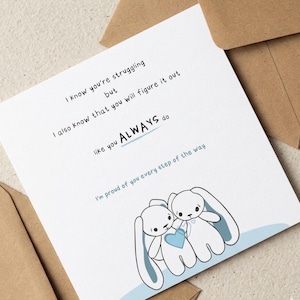 Peut inclure: Carte de v&oelig;ux blanche avec une illustration de deux lapins blancs tenant un c&oelig;ur bleu. Le texte sur la carte dit : &laquo; Je sais que vous luttez, mais je sais aussi que vous allez vous en sortir comme vous le faites TOUJOURS. Je suis fier de vous. &raquo;
