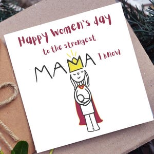 May include: A greeting card with the text "Happy Women's Day to the strongest MAMA I know." The card features a simple illustration of a woman wearing a crown and cape, holding a baby. The card is on a brown surface.