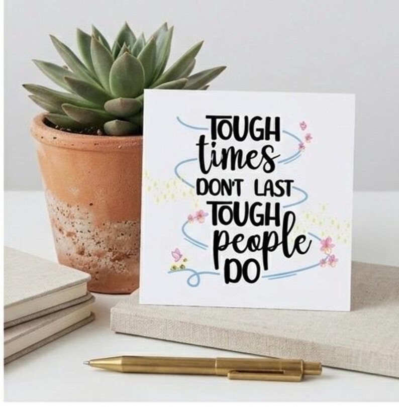 May include: A white card with the text "TOUGH times DON'T LAST TOUGH people DO" in black script, decorated with blue swirls and pink flowers. A succulent plant in a terracotta pot and a gold pen are also visible.
