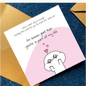Puede incluir: Una tarjeta de felicitaci&oacute;n blanca con detalles rosas, con un personaje de dibujos animados y el texto "I'm soooo glad that you're a part of my life." La tarjeta est&aacute; sobre una superficie azul, con un sobre marr&oacute;n visible.