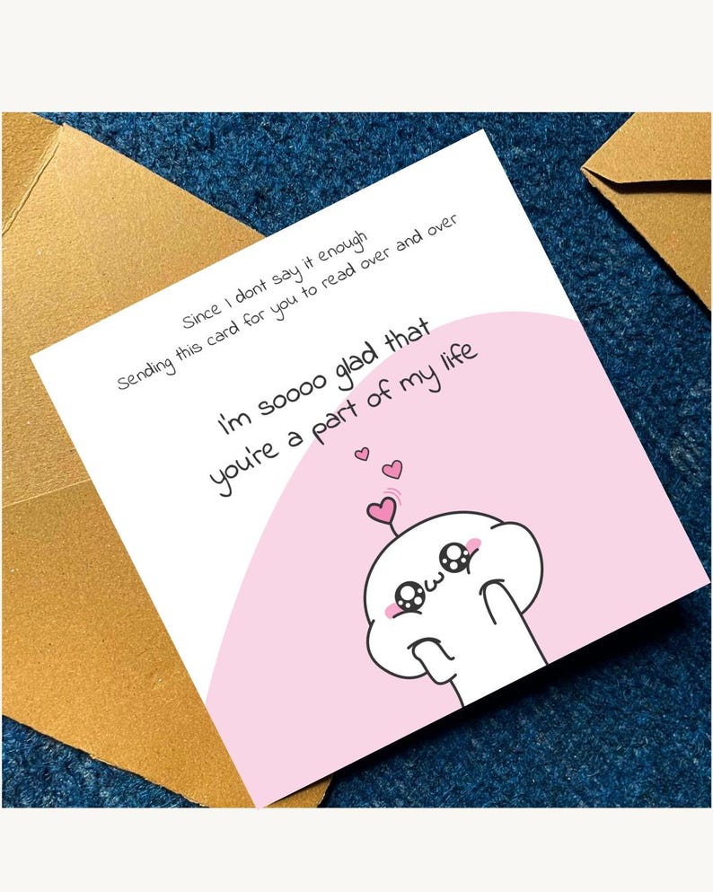 Puede incluir: Una tarjeta de felicitaci&oacute;n blanca con detalles rosas, con un personaje de dibujos animados y el texto "I'm soooo glad that you're a part of my life." La tarjeta est&aacute; sobre una superficie azul, con un sobre marr&oacute;n visible.