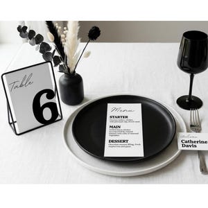 Op de afbeelding: Een monochrome tafelsetting met een zwart bord en een menukaart. Een zwart wijnglas, een tafelnummerkaart met het nummer 6 en een naambordje zijn op een wit tafelkleed geplaatst. Droogbloemen in een zwarte vaas.