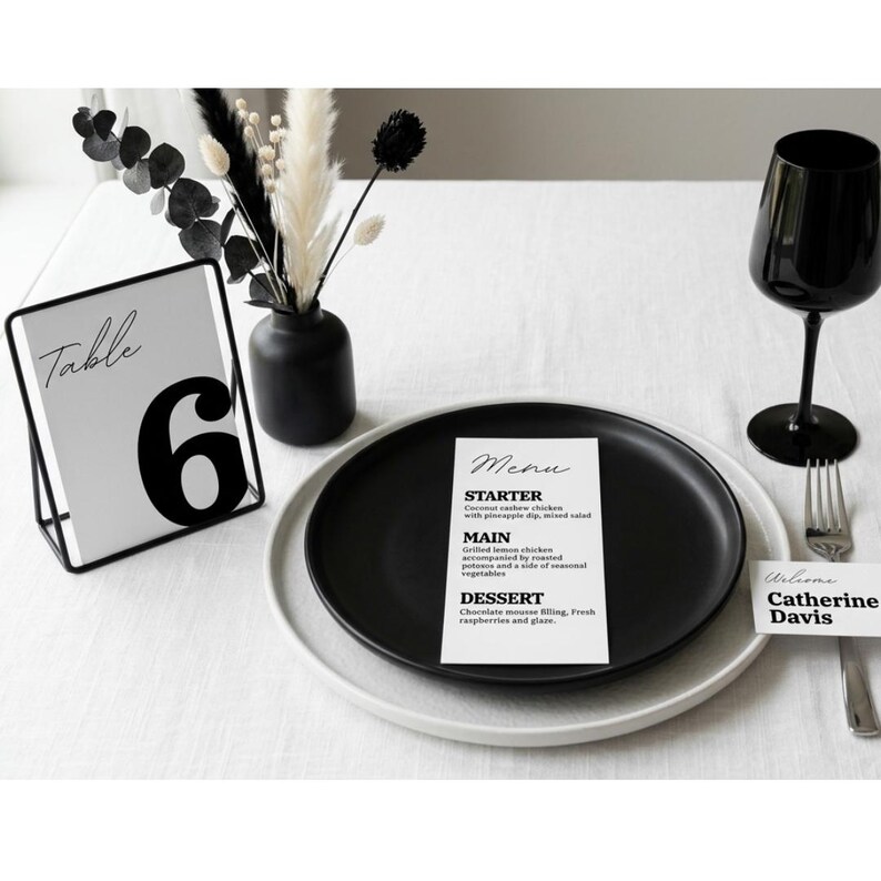 Op de afbeelding: Een monochrome tafelsetting met een zwart bord en een menukaart. Een zwart wijnglas, een tafelnummerkaart met het nummer 6 en een naambordje zijn op een wit tafelkleed geplaatst. Droogbloemen in een zwarte vaas.