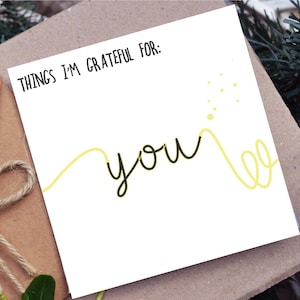 Pode incluir: Um cart&atilde;o branco com as palavras "THINGS I'M GRATEFUL FOR: YOU" em escrita preta. Um design de linha amarela acentua a palavra "you". O cart&atilde;o est&aacute; em um envelope marrom, com um la&ccedil;o de barbante e vegeta&ccedil;&atilde;o no fundo.