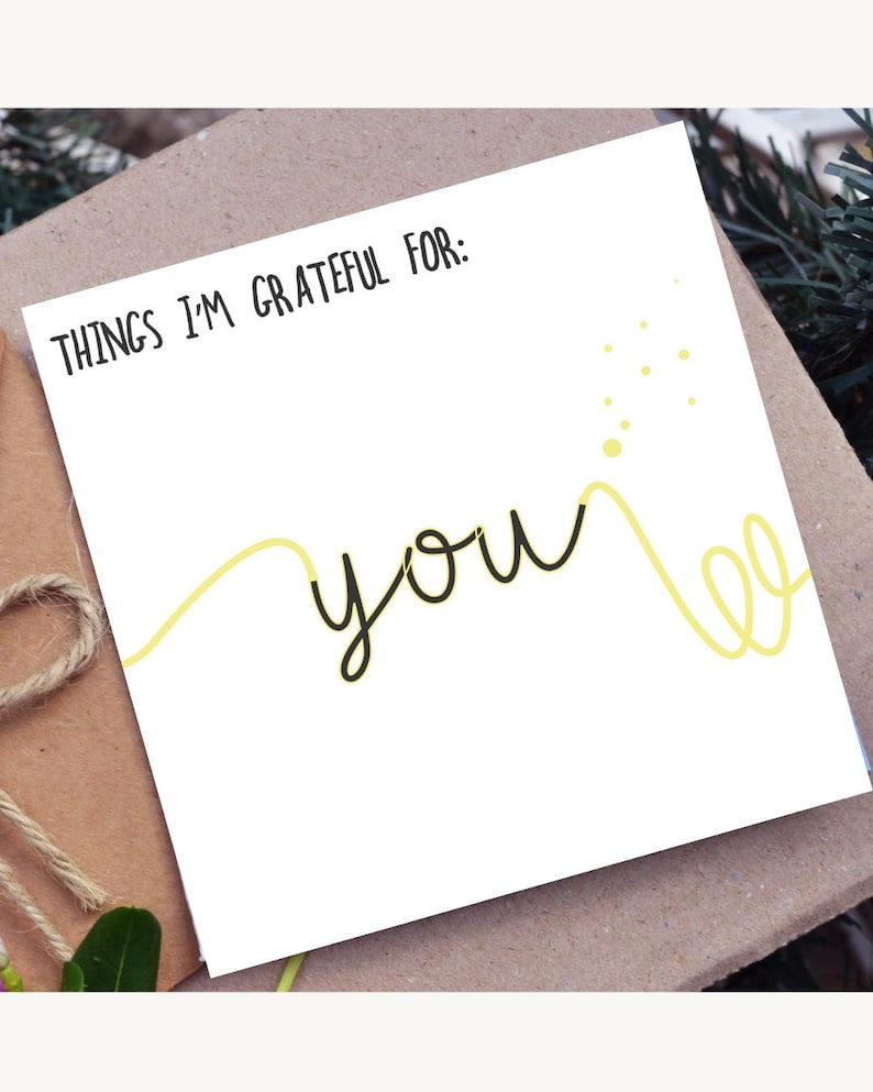 Pode incluir: Um cart&atilde;o branco com as palavras "THINGS I'M GRATEFUL FOR: YOU" em escrita preta. Um design de linha amarela acentua a palavra "you". O cart&atilde;o est&aacute; em um envelope marrom, com um la&ccedil;o de barbante e vegeta&ccedil;&atilde;o no fundo.