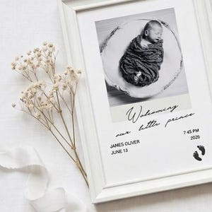May include: A white framed print featuring a black and white photograph of a swaddled newborn baby. The text reads "Welcoming our little prince", "JAMES OLIVER", "JUNE 13", "7 45 PM", and "2026", accompanied by two small black footprint icons.