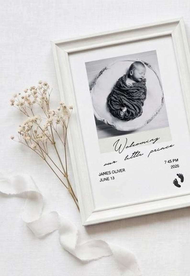 May include: A white framed print featuring a black and white photograph of a swaddled newborn baby. The text reads "Welcoming our little prince", "JAMES OLIVER", "JUNE 13", "7 45 PM", and "2026", accompanied by two small black footprint icons.