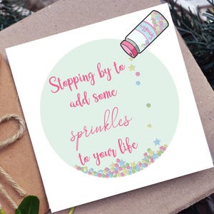 Puede incluir: Una tarjeta de felicitaci&oacute;n con el texto "Stopping by to add some sprinkles to your life" en escritura rosa. La tarjeta presenta una ilustraci&oacute;n de dibujos animados de un recipiente de chispas y chispas coloridas.
