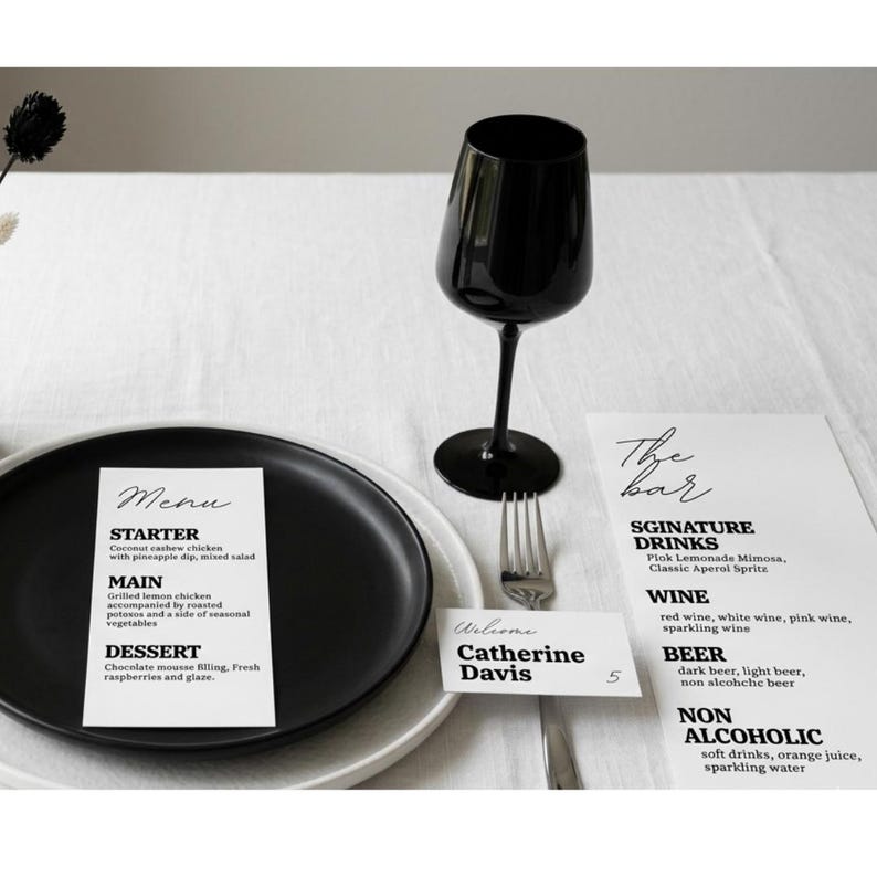 Puede incluir: Una copa de vino negra, un tenedor y un plato sobre un mantel blanco. Tambi&eacute;n hay una tarjeta de men&uacute; y una carta de bebidas. La tarjeta del men&uacute; enumera un entrante, un plato principal y un postre. La carta de bebidas enumera bebidas de autor, vino, cerveza y bebidas sin alcohol.