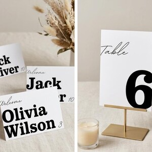 May include: White table cards with black text, including names and numbers. One card reads "Table 6" on a gold stand. Other cards display names like "Jack Oliver" and "Olivia Wilson". A lit candle sits nearby.
