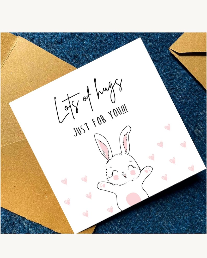 May include: A white greeting card with the text "Lots of hugs JUST FOR YOU!!!" features a smiling cartoon bunny with outstretched arms and pink hearts. The card is accompanied by a gold envelope, set against a dark blue textured background.