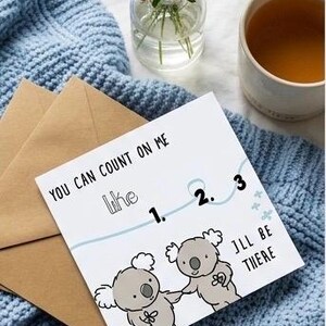 May include: A greeting card with the text "You can count on me like 1. 2. 3. I'll be there" and koala bear illustrations. The card is on a blue knitted blanket with a brown envelope. A vase of flowers and a cup of tea are also present.