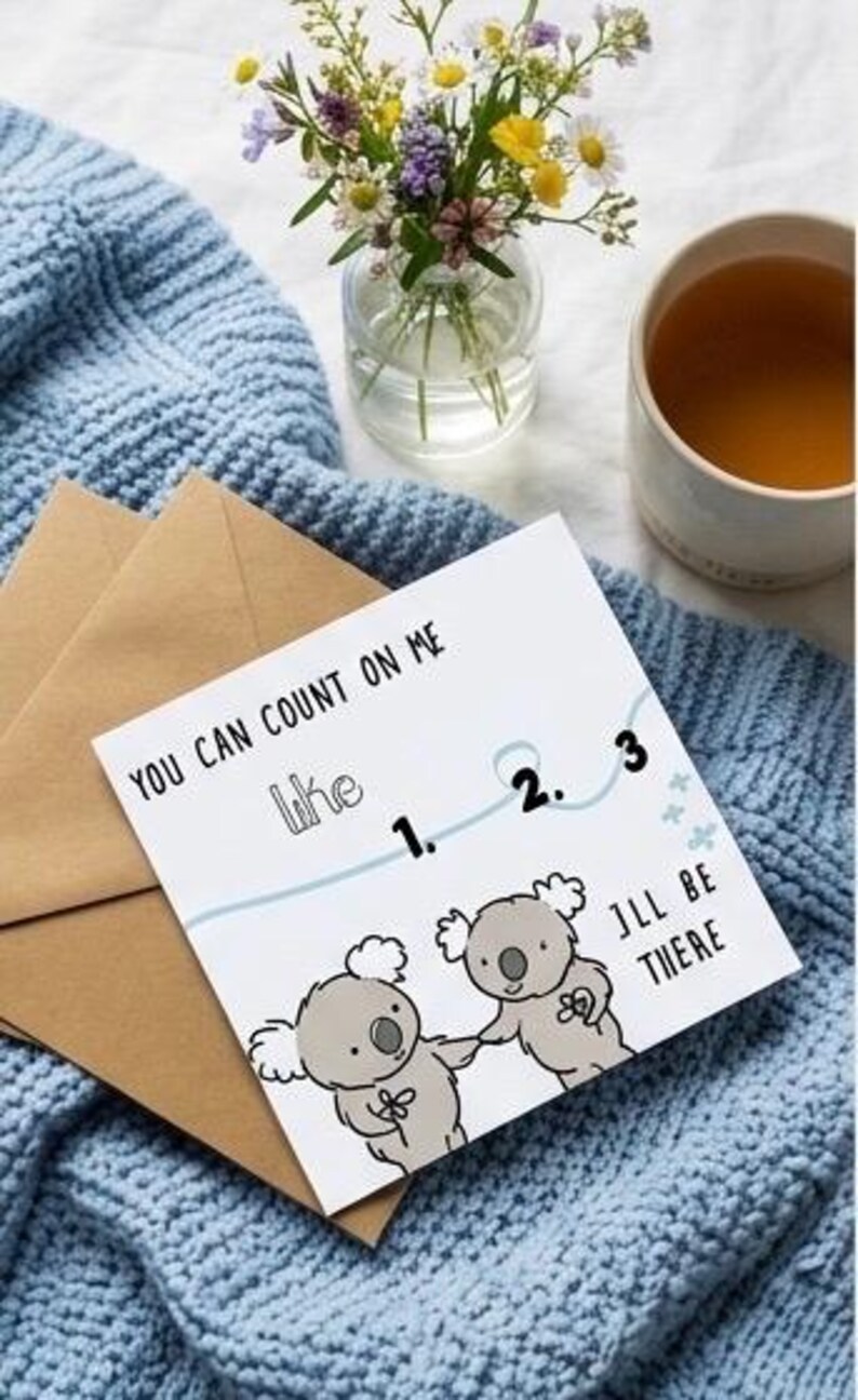 May include: A greeting card with the text "You can count on me like 1. 2. 3. I'll be there" and koala bear illustrations. The card is on a blue knitted blanket with a brown envelope. A vase of flowers and a cup of tea are also present.