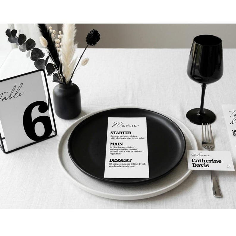May include: A table setting with a black plate, a menu card, and a black wine glass. The menu card lists a starter, main course, and dessert. A table number 6 card and a welcome card are also present.