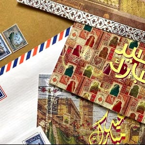 May include: A collection of stationery featuring a white Air Mail envelope with red and blue stripes, postage stamps, and greeting cards with Arabic script and decorative patterns. The items are displayed on a brown surface.