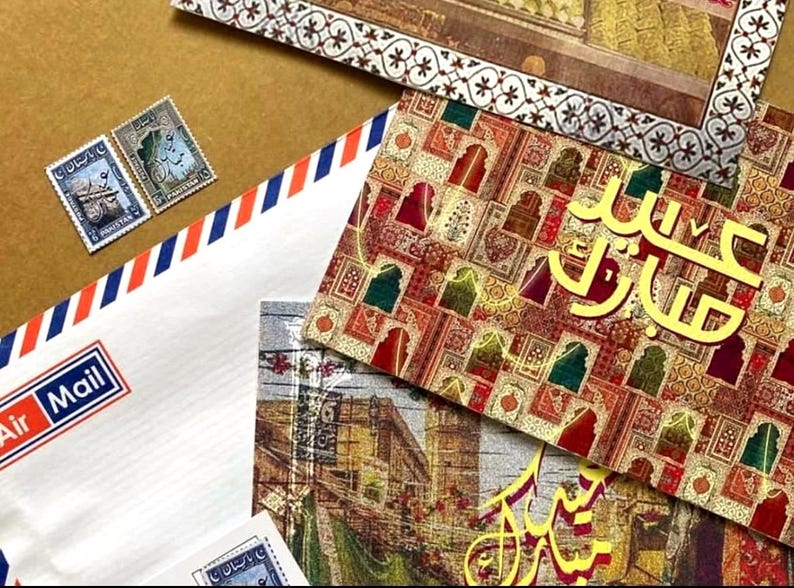 May include: A collection of stationery featuring a white Air Mail envelope with red and blue stripes, postage stamps, and greeting cards with Arabic script and decorative patterns. The items are displayed on a brown surface.