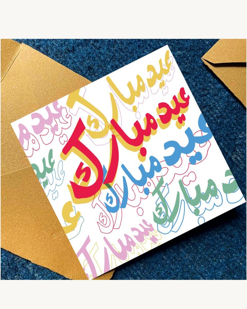 Peut inclure: Une carte de v&oelig;ux blanche avec une calligraphie arabe color&eacute;e disant "Eid Mubarak" en rouge, jaune, bleu et vert. La carte est pos&eacute;e sur une surface bleu fonc&eacute; avec une enveloppe dor&eacute;e.