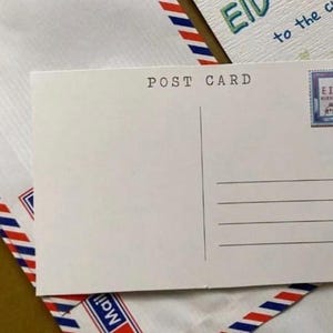 May include: A white postcard with the words "POST CARD" printed at the top. The postcard has lines for writing and a stamp with the word "EID" on it. The postcard is on top of an envelope with red, white, and blue stripes.