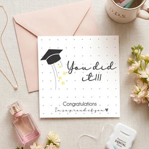 Peut inclure: Une carte de v&oelig;ux carr&eacute;e blanche avec une ic&ocirc;ne de toque de dipl&ocirc;m&eacute; noire, des &eacute;toiles jaunes et le texte "You did it!!! Congratulations I'm so proud of you" avec un c&oelig;ur, sur un fond clair avec une enveloppe rose et des accessoires.