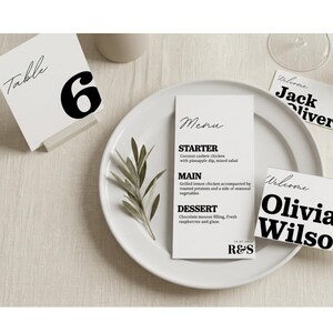 Op de afbeelding: Een witte placering met een menukaart, tafelnummer en naambordjes. De menukaart bevat een voorgerecht, hoofdgerecht en dessert. Het tafelnummer is "6". De naambordjes zeggen "Welcome" en bevatten namen.
