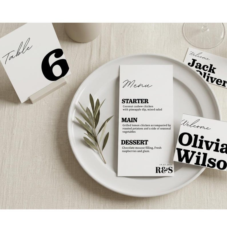Op de afbeelding: Een witte placering met een menukaart, tafelnummer en naambordjes. De menukaart bevat een voorgerecht, hoofdgerecht en dessert. Het tafelnummer is "6". De naambordjes zeggen "Welcome" en bevatten namen.