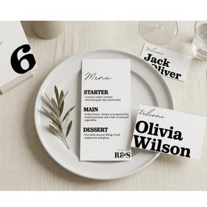 Op de afbeelding: Een witte plaat met een menukaart, een tafelnummer en twee naamkaarten. De menukaart bevat een voorgerecht, hoofdgerecht en dessert. De naamkaarten zeggen "Welkom" en de namen "Jack Oliver" en "Olivia Wilson".
