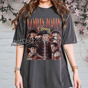 May include: Dark gray t-shirt featuring a graphic of Lord John Grey with text. The graphic includes images of the character in historical attire. The text "LORD JOHN Grey" is in a distressed, vintage style.