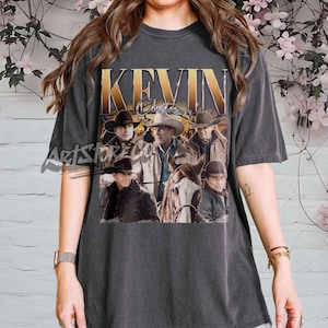 Könnte beinhalten: Dunkelgraues T-Shirt mit einem Grafikdesign im Vintage-Stil. Die Grafik zeigt den Text "KEVIN Costner" in Gold und ein Sepia-Bild von Männern in Cowboyhüten und Jacken, mit einem Pferd. Retro-Design.