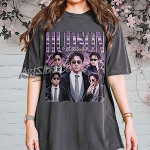 Puede incluir: Camiseta gris oscuro con un gráfico de un hombre con traje y gafas de sol, con el nombre "HUDSON" en texto morado. La camiseta tiene un aspecto vintage y desgastado.